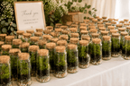 A table covered with many small glass jars containing green plants and pebbles, arranged in rows on a white cloth. A framed sign reading 'Thank you for being part of our special day' stands among the jars, with floral arrangements and a softly lit indoor setting in the background.